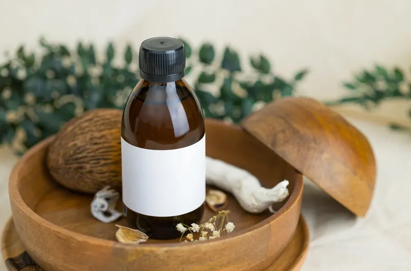 A brown glass bottle with a blank label placed on a wooden plate surrounded by botanical elements, ideal for cosmetic and natural product themes