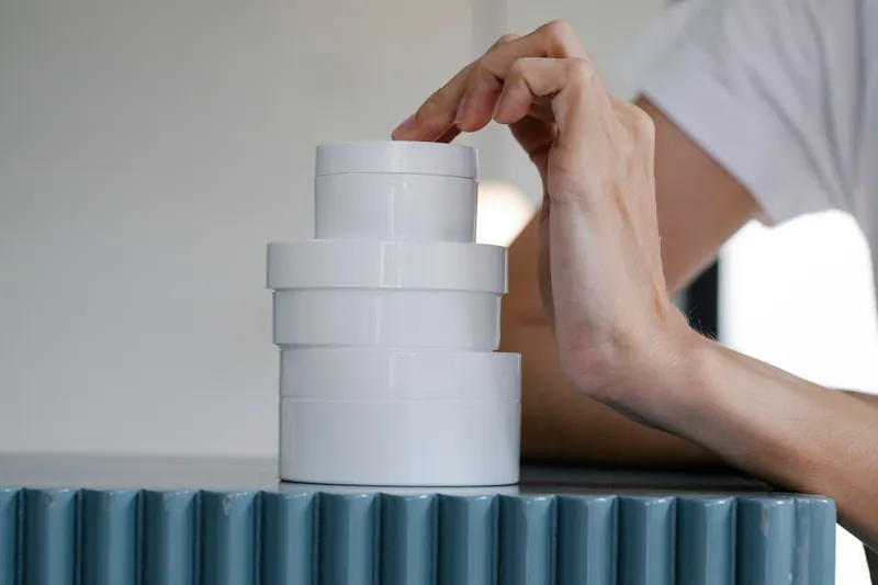 Crop anonymous female in t shirt leaning on table and touching stacked white jars of skin care products