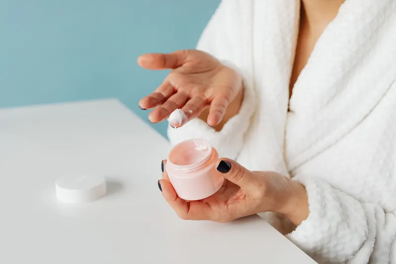 A woman in robe applying skincare cream from an open jar, highlighting moisturizing routine