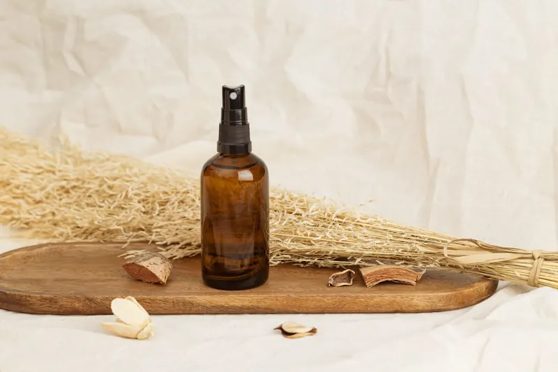 Amber glass spray bottle placed on a wooden tray with dried plants, suggesting natural cosmetics