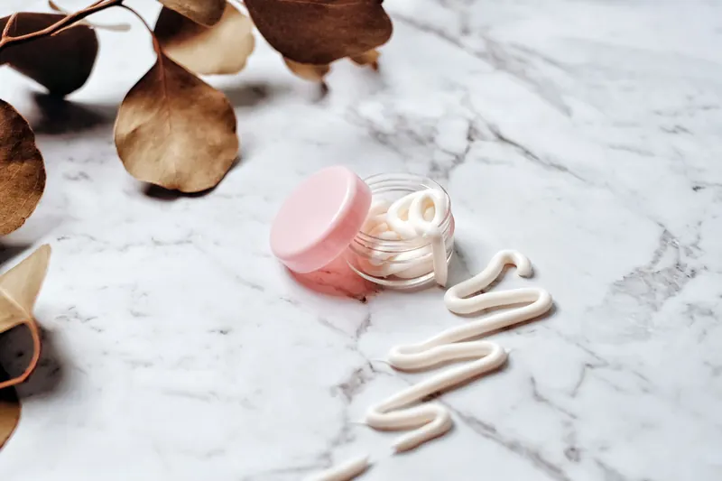 Close-up of skincare cream in a pink glass jar on a marble surface surrounded by leaves, ideal for beauty and wellness themes